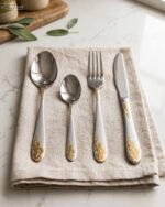 24-piece silver stainless steel cutlery set with gold floral embossed handles displayed on a linen napkin.