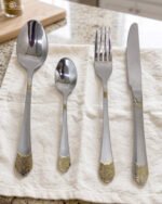 24-piece polished stainless steel cutlery set with gold filigree handle accents arranged on a beige cloth.