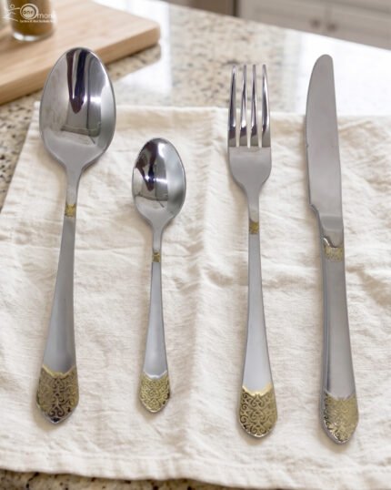 24-piece polished stainless steel cutlery set with gold filigree handle accents arranged on a beige cloth.