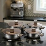 Four stainless steel double-handle woks with glass lids in 24cm, 26cm, 28cm, and 30cm sizes displayed on a dark kitchen countertop.