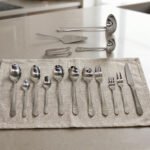 A Diverse Arrangement of Polished Silver-Toned Cutlery on a Textured Linen Mat, Featuring 12-Person Service and Loose Ladles, a Cake Server, and Tongs in the Background.