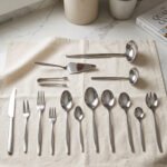 72-piece polished stainless steel cutlery set arranged on a cloth napkin featuring place settings, ladles, cake server, and tongs.