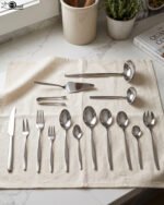 72-piece polished stainless steel cutlery set arranged on a cloth napkin featuring place settings, ladles, cake server, and tongs.