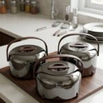 Three modern mirror-finish stainless steel insulated hot pots with wooden arch handles on a kitchen countertop.