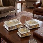 Three silver textured decorative serving dishes with gold floral accents and overhead arched handles on a wooden coffee table.