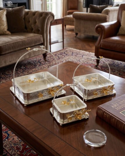 Three silver textured decorative serving dishes with gold floral accents and overhead arched handles on a wooden coffee table.