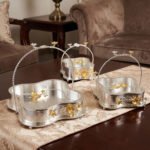 Three-piece silver hammered decorative bowl set with gold floral accents and overhead handles arranged on a wooden table.