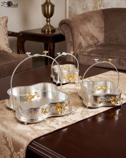 Three-piece silver hammered decorative bowl set with gold floral accents and overhead handles arranged on a wooden table.