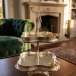 Two-tier silver hammered decorative serving tray with gold floral accents resting on a wooden coffee table in an elegant living room setting.