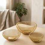 Set of three gold mesh decorative wire bowls sitting on a light wooden table in a brightly lit modern living room setup.