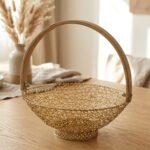 Gold decorative metal mesh basket with a curved wooden handle placed on a wooden table in a bright modern dining room