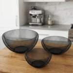 Set of three round black wire mesh decorative metal baskets displayed on a wooden kitchen countertop.