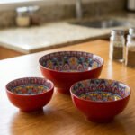 A set of three decorative ceramic bowls in a kitchen. They have rich red exterior glazes and detailed, traditional hand-painted Moroccan-style patterns in blue, yellow, and red on the inside, sitting on a wooden counter next to spice jars.