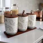 Three white ceramic kitchen canisters with gray leaf patterns, wooden lids with loop handles, and a wooden tray displayed on a white kitchen countertop.
