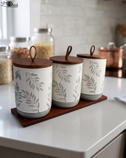 Three white ceramic kitchen canisters with gray leaf patterns, wooden lids with loop handles, and a wooden tray displayed on a white kitchen countertop.