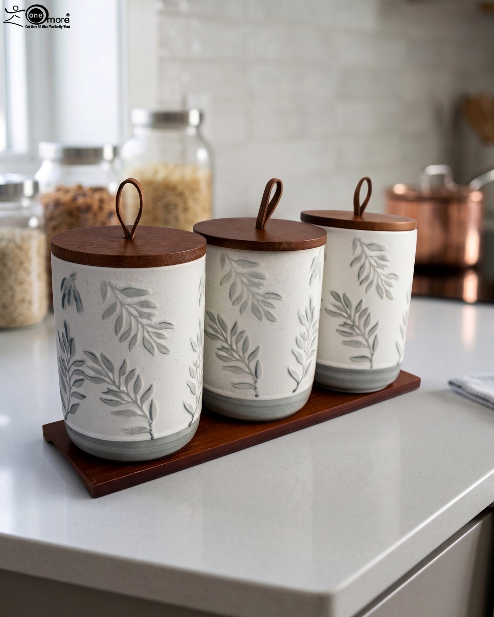 1 Three white ceramic kitchen canisters with gray leaf patterns, wooden lids with loop handles, and a wooden tray displayed on a white kitchen countertop.