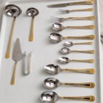 72-piece stainless steel cutlery set with gold trim handles and specialty serving utensils displayed on a white tray.