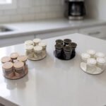 Four modern 6-piece revolving glass spice jar sets with gold trim lids displayed on a white kitchen countertop in beige, cream, black, and white colors.