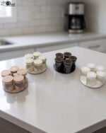 Four modern 6-piece revolving glass spice jar sets with gold trim lids displayed on a white kitchen countertop in beige, cream, black, and white colors.