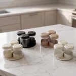 Four modern glass spice jar sets in black, tan, cream, and off-white with gold lid rings arranged on a marble kitchen countertop.