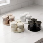 Four sets of ONE MORE 3-piece glass spice jars with trays sitting on a white marble countertop in beige, off-white, white, and black colors.