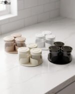 Four sets of ONE MORE 3-piece glass spice jars with trays sitting on a white marble countertop in beige, off-white, white, and black colors.