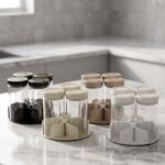 Four-piece clear glass spice jar sets with airtight lids displayed on rotating organizer trays in black, beige, tan, and white on a marble kitchen countertop.