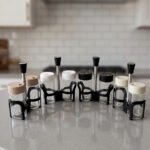 A close-up shot of an 8-piece glass spice rack set with assorted color lids and black handles arranged on a grey kitchen countertop with a blurred backsplash.