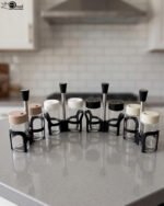 A close-up shot of an 8-piece glass spice rack set with assorted color lids and black handles arranged on a grey kitchen countertop with a blurred backsplash.