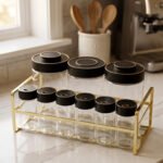 9-piece luxury glass spice jars with black gold-accented lids stored on a metallic gold wire rack on a marble kitchen countertop.