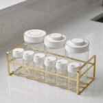 9-piece clear glass spice jar set with white and gold airtight lids arranged on a gold metal rack on a kitchen countertop.