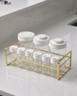 9-piece clear glass spice jar set with white and gold airtight lids arranged on a gold metal rack on a kitchen countertop.