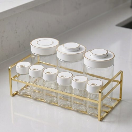 9-piece clear glass spice jar set with white and gold airtight lids arranged on a gold metal rack on a kitchen countertop.