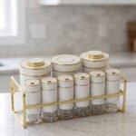 9-piece white ceramic and clear glass spice jar set with gold accents arranged in a gold metal rack on a marble countertop