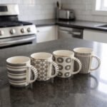 Set of 4 ceramic coffee mugs with charcoal grey geometric and striped patterns on a dark granite kitchen island counter.