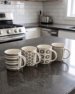 Set of 4 ceramic coffee mugs with charcoal grey geometric and striped patterns on a dark granite kitchen island counter.