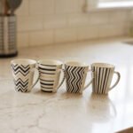Set of four modern ceramic coffee mugs featuring black and white geometric, chevron, and striped patterns with gold rim accents on a marble countertop.