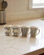 Set of four modern ceramic coffee mugs featuring black and white geometric, chevron, and striped patterns with gold rim accents on a marble countertop.