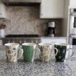 Four ceramic coffee cups featuring green, yellow, and beige geometric and botanical patterns lined up on a granite kitchen countertop