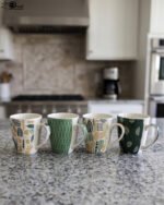 Four ceramic coffee cups featuring green, yellow, and beige geometric and botanical patterns lined up on a granite kitchen countertop