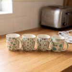 Four Onemore ceramic coffee mugs with teal and grey geometric patterns arranged on a wooden kitchen countertop.