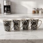 Set of four modern ceramic coffee mugs featuring black and white giraffe, zebra, and leopard prints with gold trim sitting on a marble kitchen counter.