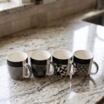 Set of four modern ceramic coffee mugs featuring black and white geometric patterns lined up on a granite countertop.
