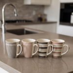 Four ceramic coffee cups with vertical and horizontal striped patterns lined up on a kitchen counter