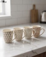 Set of four cream ceramic coffee mugs with metallic heart patterns and gold rims displayed on a marble kitchen countertop