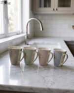 Four ceramic coffee mugs in beige, mint green, blush pink, and light blue with floral geometric patterns displayed on a marble countertop.
