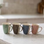 4-piece ceramic coffee mug set with gold star patterns in sage green, brown, navy blue, and blush pink on a white marble countertop.