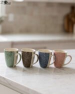 4-piece ceramic coffee mug set with gold star patterns in sage green, brown, navy blue, and blush pink on a white marble countertop.