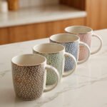 Four ceramic coffee mugs with pastel floral patterns in grey, green, blue, and pink featuring gold rims on a marble kitchen countertop.