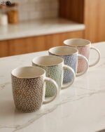 Four ceramic coffee mugs with pastel floral patterns in grey, green, blue, and pink featuring gold rims on a marble kitchen countertop.